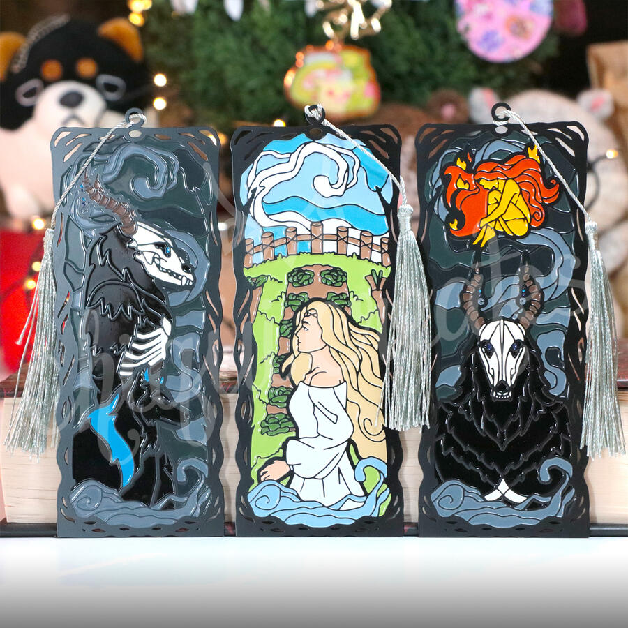 Three metal bookmarks with characters from A Soul to Keep. From left: Orpheus and his wolf, Rhea in front of her farm, and Orpheus with Rhea's flame form floating above his horns.