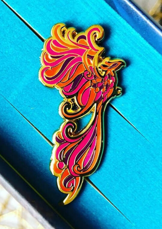 Gold enamel pin of a flowy phoenix design with orange, magenta, and dark purple feathers.